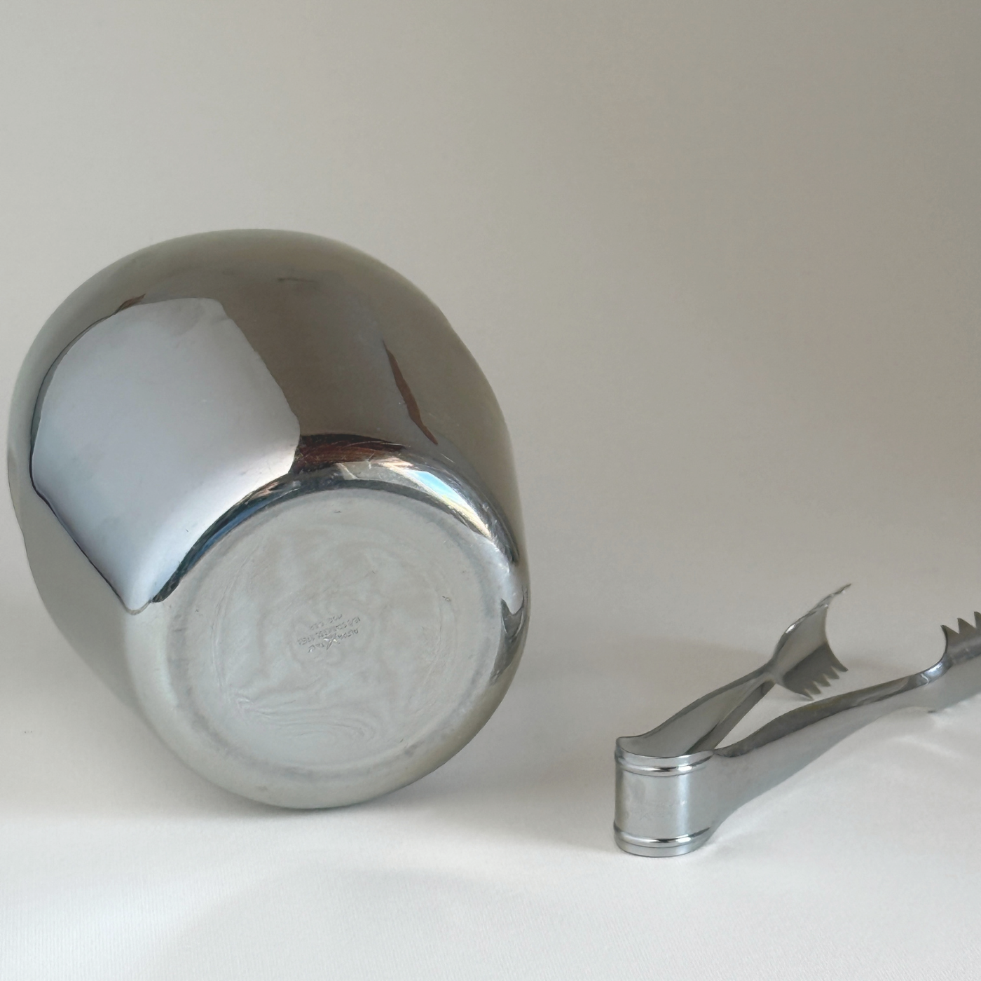 Alessi 871 Ice Bucket in mirror-polished stainless steel with smooth cylindrical form, shown with matching ice tongs on white surface.