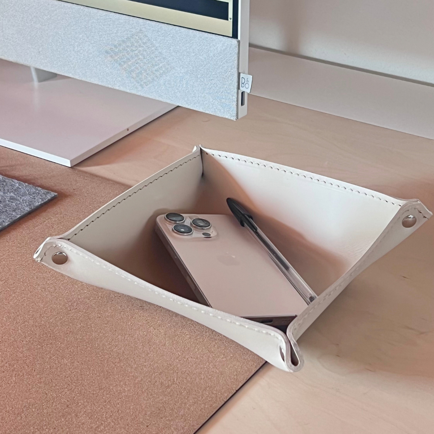 A cream-colored leather desk organizer tray with metal grommets, shown on a light wood desk holding a smartphone and a pen, with a monitor base visible in the background.
