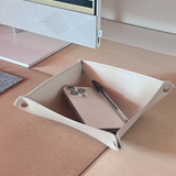 A cream-colored leather desk organizer tray with metal grommets, shown on a light wood desk holding a smartphone and a pen, with a monitor base visible in the background.