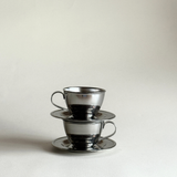 Two silver teacups with saucers on a light gray background P&B nanni