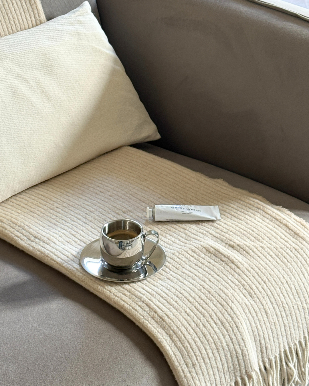 Silver imco coffee cup and saucer on a beige cashmere blanket with a white pillow on a grey couch byredo gipsy water white hand cream. 