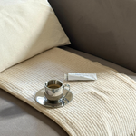 Silver imco coffee cup and saucer on a beige cashmere blanket with a white pillow on a grey couch byredo gipsy water white hand cream. 