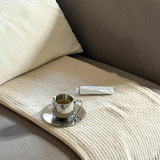Silver imco coffee cup and saucer on a beige cashmere blanket with a white pillow on a grey couch byredo gipsy water white hand cream. 