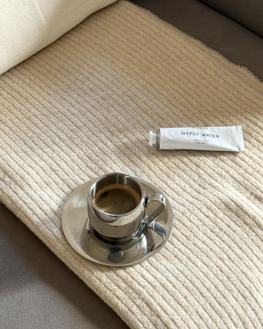 Silver imco coffee cup and saucer on a beige cashmere blanket with byredo gipsy water white hand cream. 