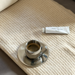 Silver imco coffee cup and saucer on a beige cashmere blanket with byredo gipsy water white hand cream. 