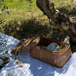Wicker picnic basket with items on a white blanket under an olive tree