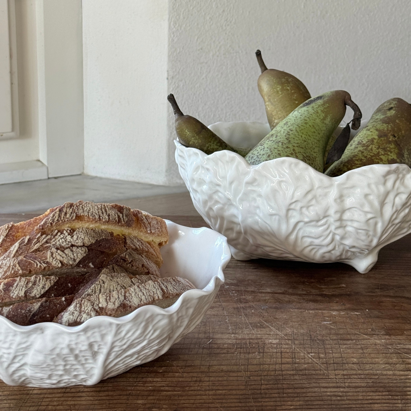 Two white ceramic bowls (wedgwood country ware and spode) with bread and pears on a wooden surface.